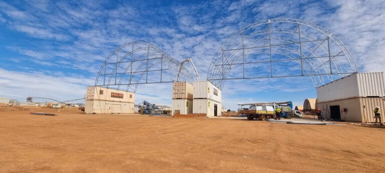 Steel frame dome shelter being installed atop stacked shipping containers on red dirt site