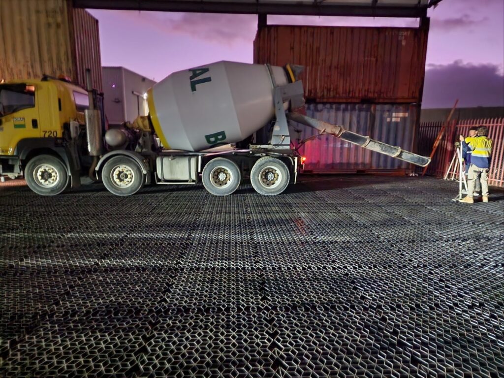 Cement truck pouring concrete over plastic grid base framed by shipping containers